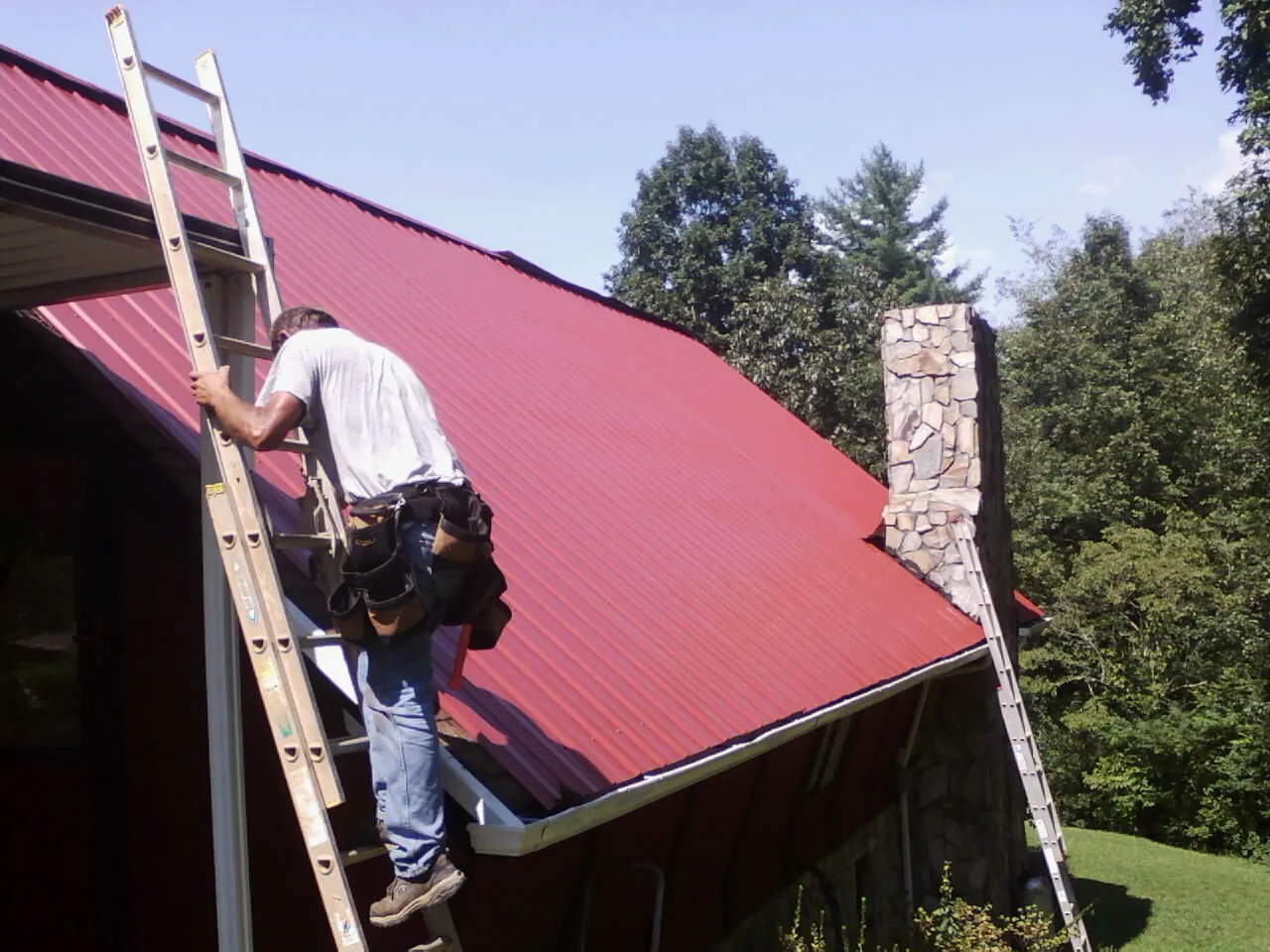 Roofer on a ladder beside a red metal roof and stone chimney — Soffit & Fascia Repair by Roofing Brothers in Waukomis